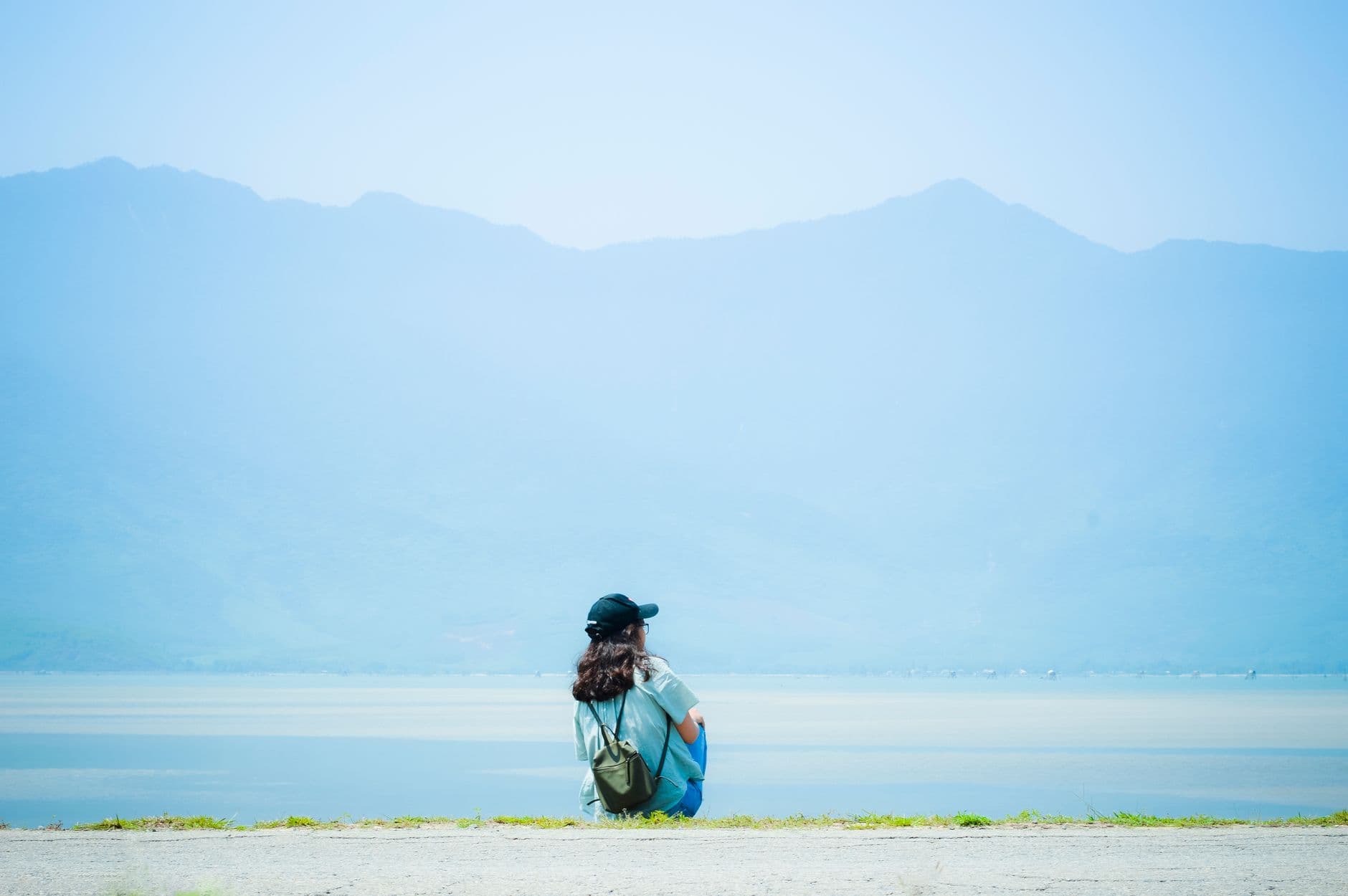 A person sitting quietly outdoors, looking out over a calm lake and hills