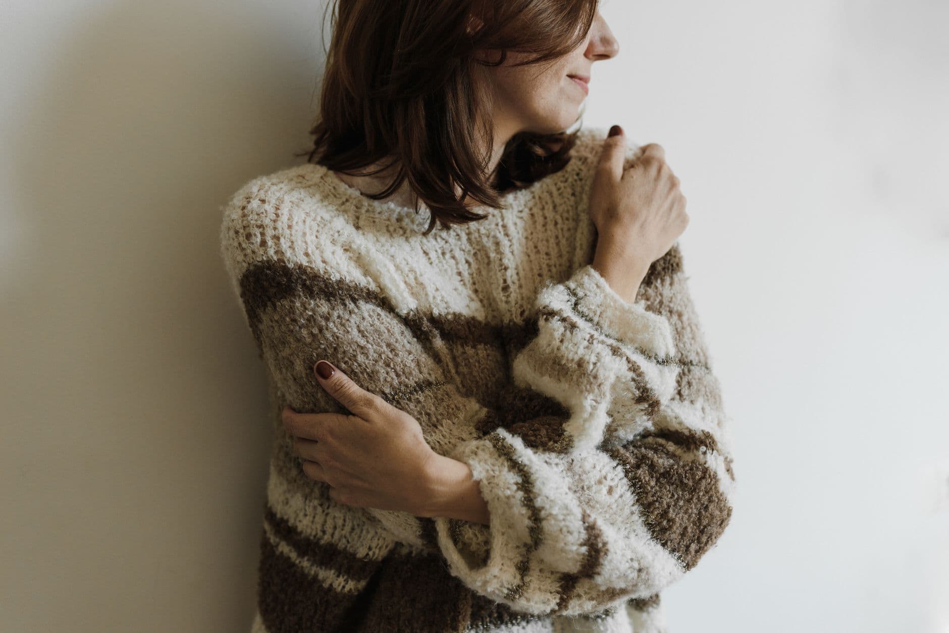 A woman in a soft knitted jumper gently hugging herself, smiling, against a pale wall